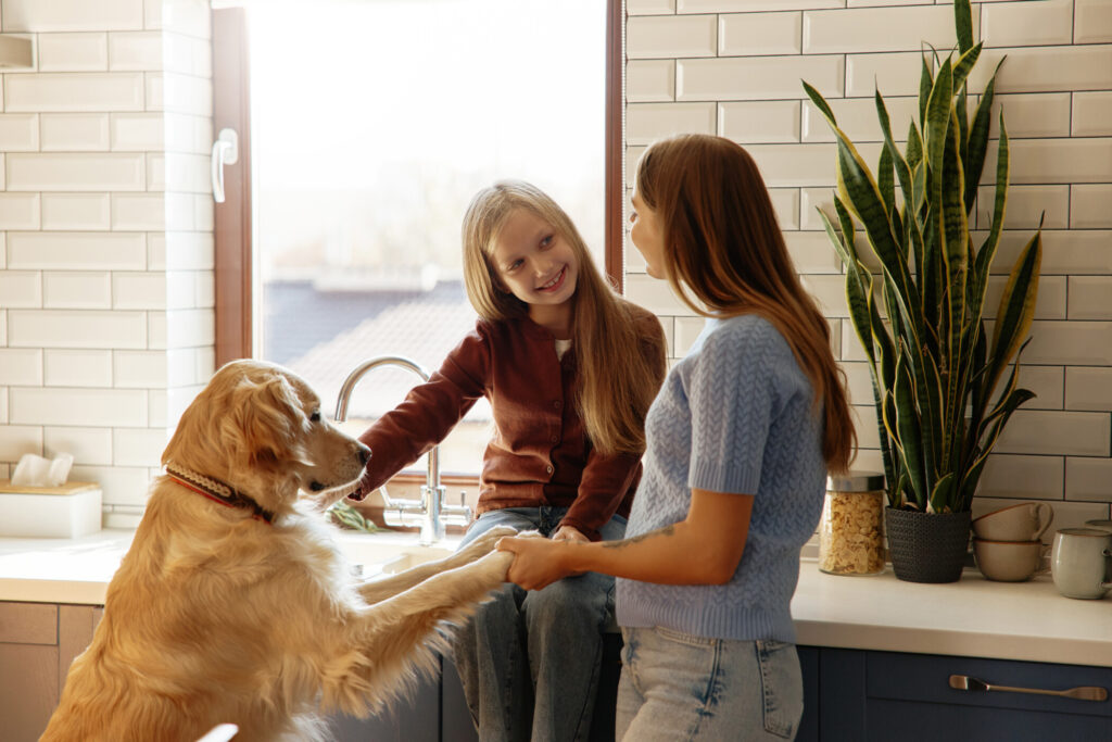 Smiling girl sits on a kitchen countertop near a sunny window, holding a smartphone. Bright cozy kitchen interior with plants, juice, and coffee machine. Concept: childhood, morning routine, tech.